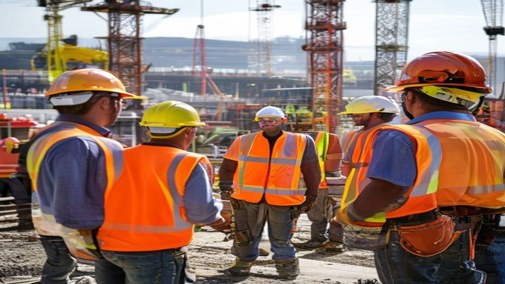 Morning safety toolbox talk on active construction site, diverse team of workers in bright orange PPE safety vests and yellow hard hats gathered around a site supervisor, steel structure and tower crane visible in background, professional documentary photography style